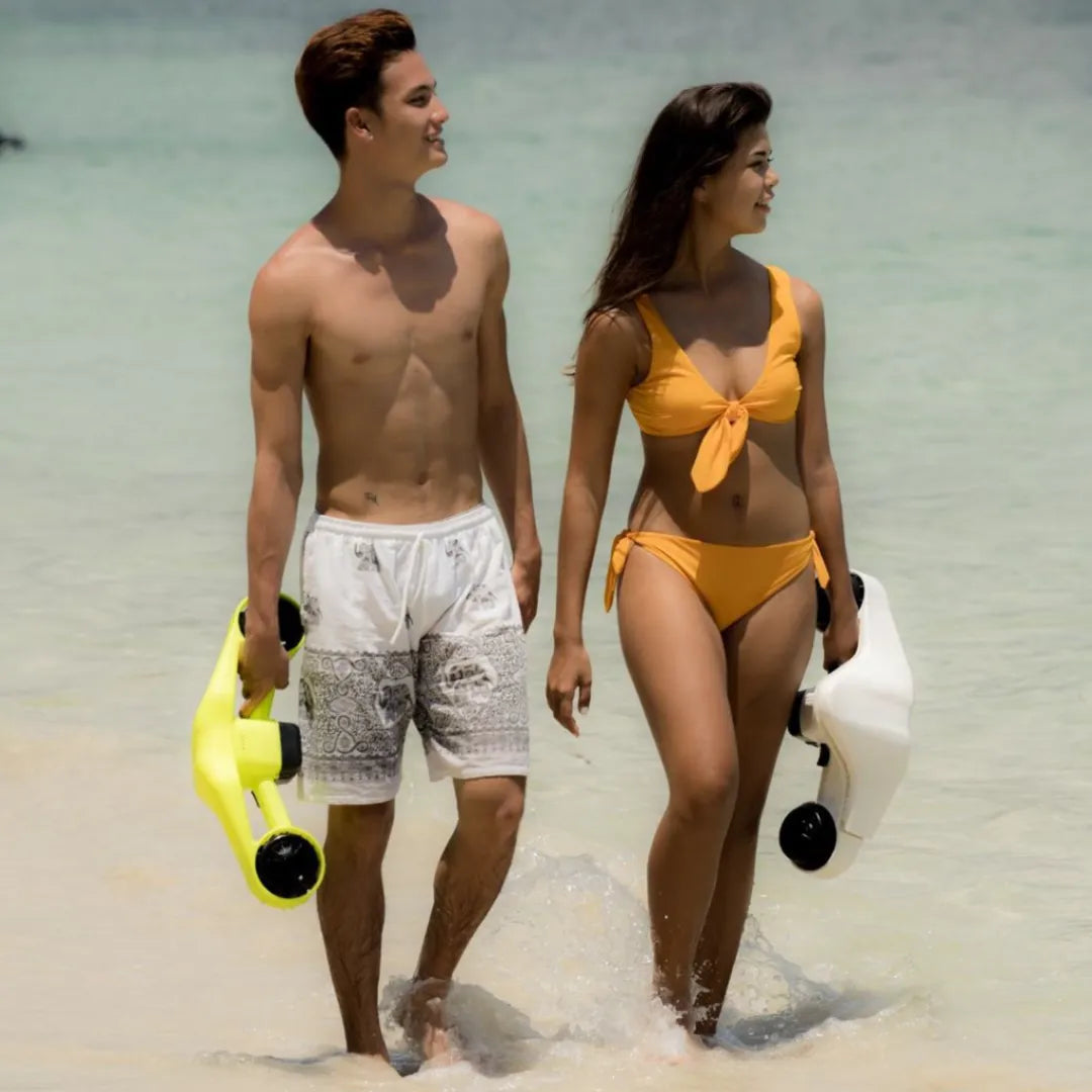 The product is an Electric Water Scooter by Asiwo, model Manta. The image shows two young people walking on the beach, dressed in swimwear. Each person is holding one of the scooters, one yellow and one white, suggesting active time in the water. Both individuals are smiling and enjoying the sunny weather.