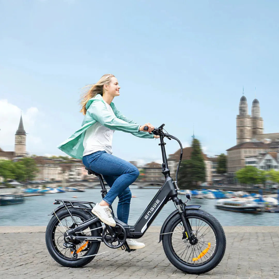Na sliki je električno kolo znamke Engwe, model L20 3.0 Pro. Prikazuje mlado žensko, ki vozi kolo ob vodi, v ozadju pa so vidni mestni arhitekturni elementi in čolni. Kolo ima kompaktno zasnovo, široke pnevmatike in je primerno za mestno vožnjo.