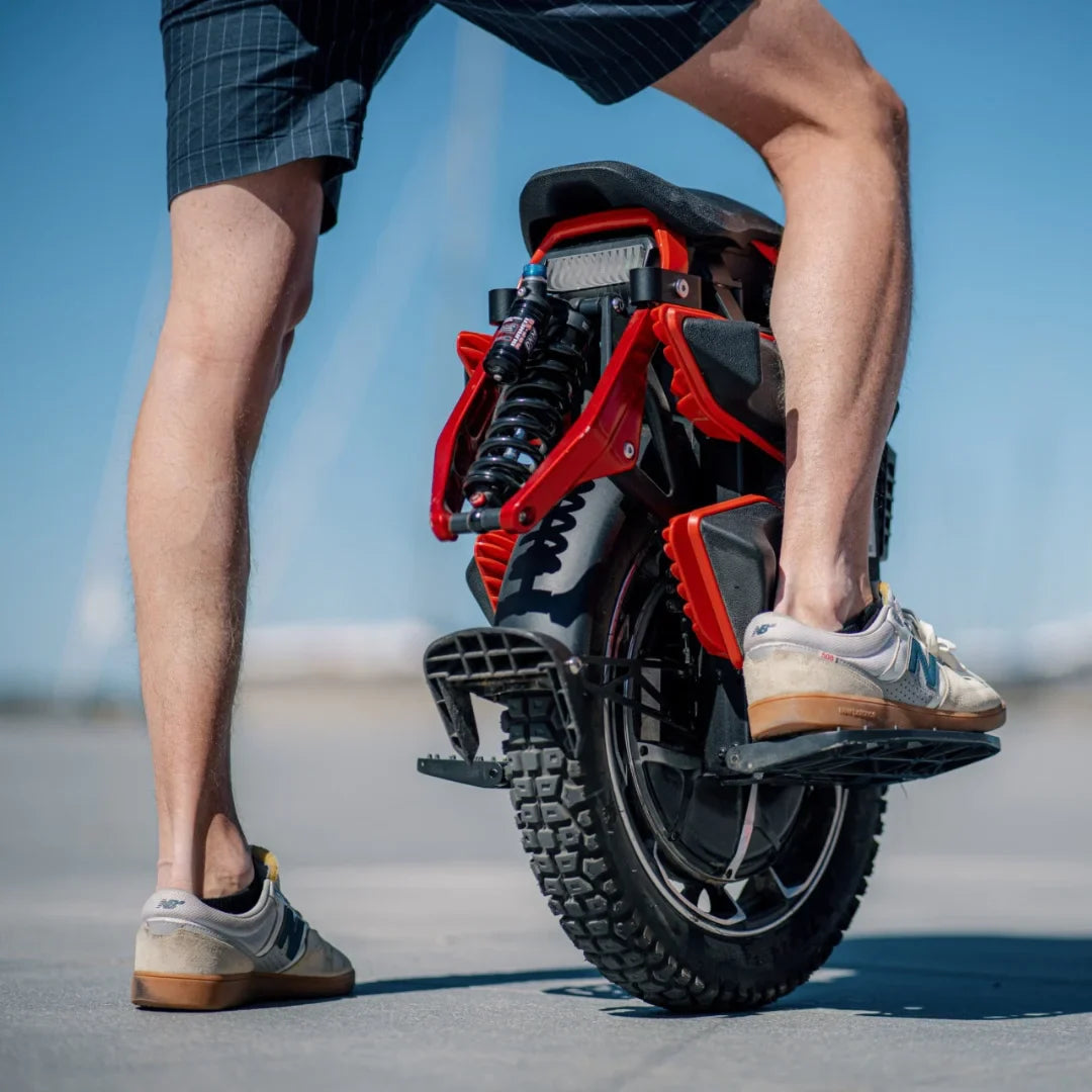 The product is an electric unicycle, KingSong model S22 Pro. The image shows the rear of the unicycle, which features a robust tire and suspension. The person standing next to it is wearing shorts and athletic shoes, suggesting a relaxed and active use of this mode of transport. In the background, there's a clear sky, creating a pleasant atmosphere.
