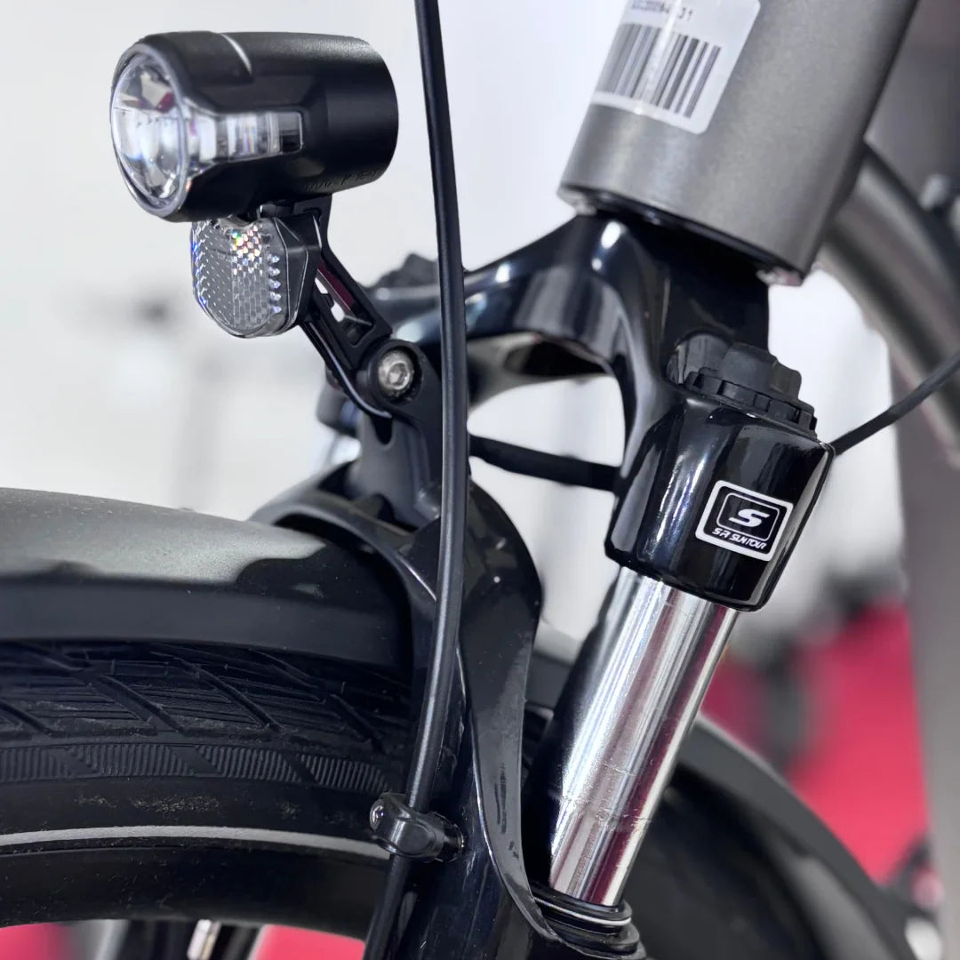 The product pictured is an Electric Bike from the brand Malaguti, model Bolonina. The image shows a part of the front suspension, including the headlight and a portion of the front fork. A tire with a tread suitable for various terrains is also visible. The bike is designed for comfortable and safe riding.