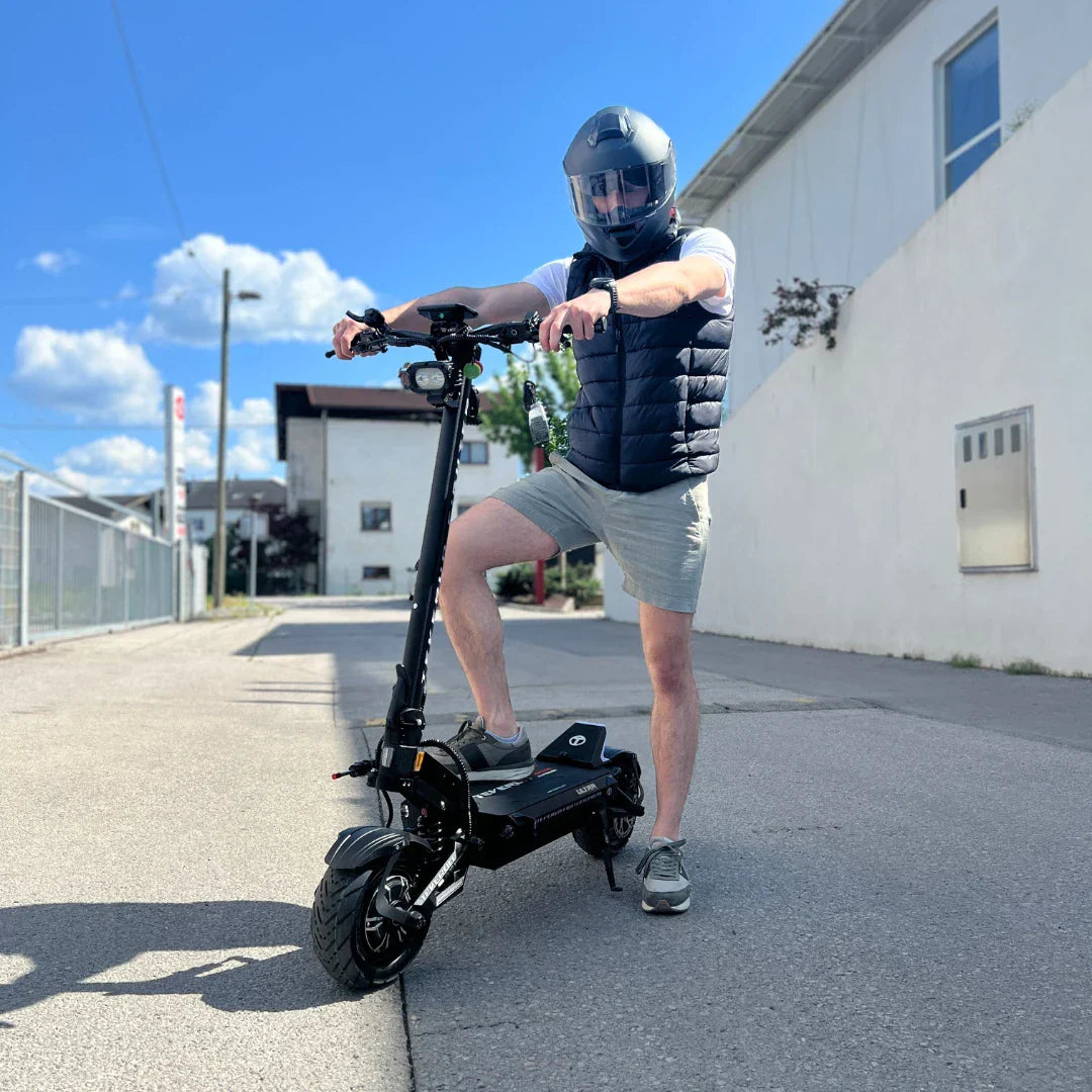 Na sliki je električni skiro, model Teverun Fighter Supreme 7260 Ultra. Izdelek je črne barve in ima robusten okvir ter široke pnevmatike, kar nakazuje na dobro stabilnost in zmogljivost. Oseba na skiroju nosi čelado in športna oblačila, kar kaže na zavedanje o varnosti pri vožnji. Ozadje prikazuje mestno okolje s stavbami in modrim nebom.