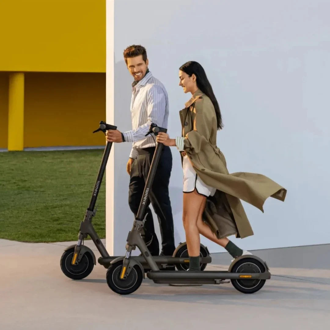 The image shows an electric scooter, model Xiaomi 5 Max GL. Two people, a man and a woman, are walking and holding scooters, which are in a neutral gray color. Both people are dressed in casual clothes, and the surroundings are modern and bright, creating a pleasant atmosphere.