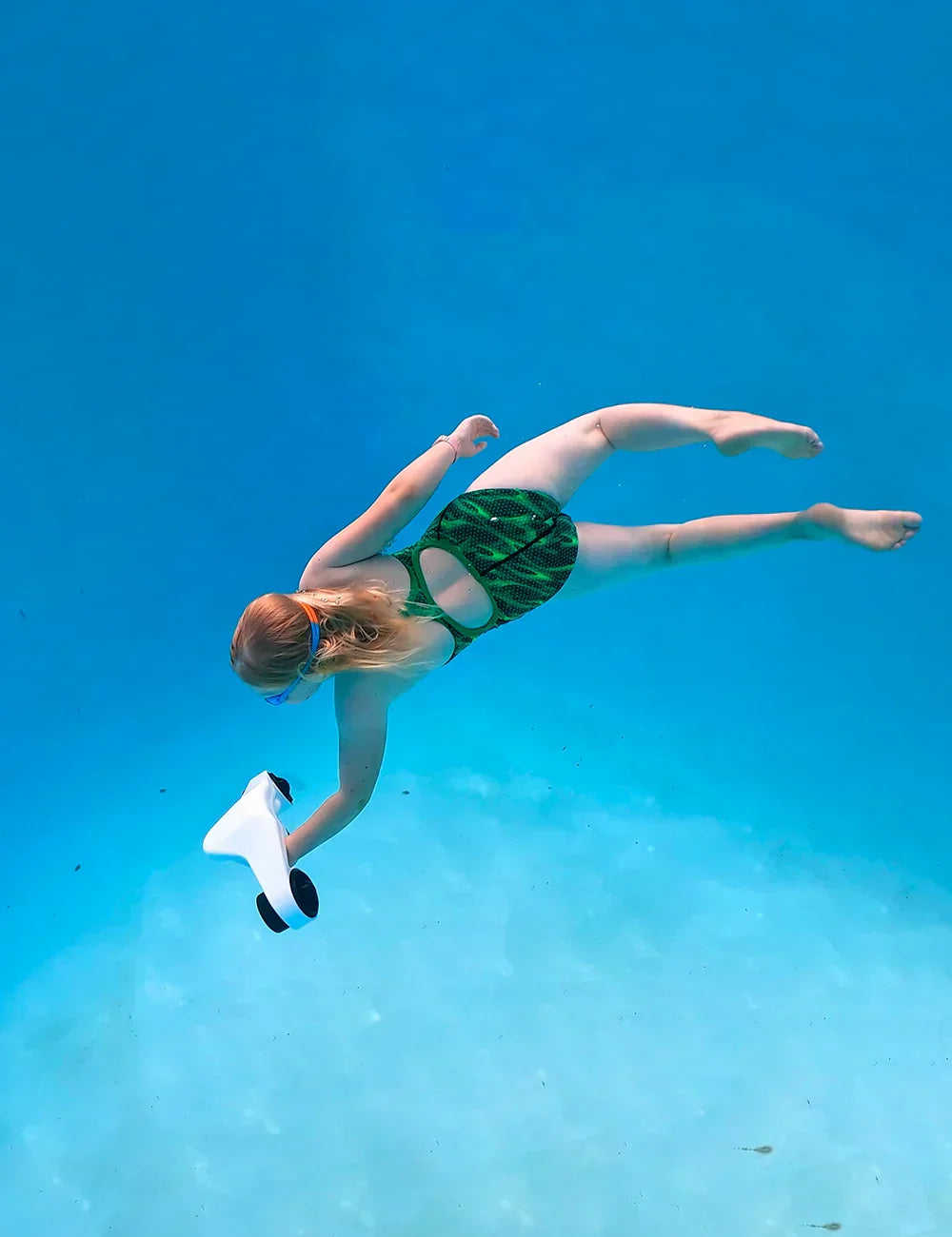 The product in the picture is an electric water scooter, model Asiwow Manta. The picture shows a girl swimming underwater and holding the scooter, dressed in a green patterned swimsuit. The background is clear blue water, creating a pleasant underwater atmosphere.
