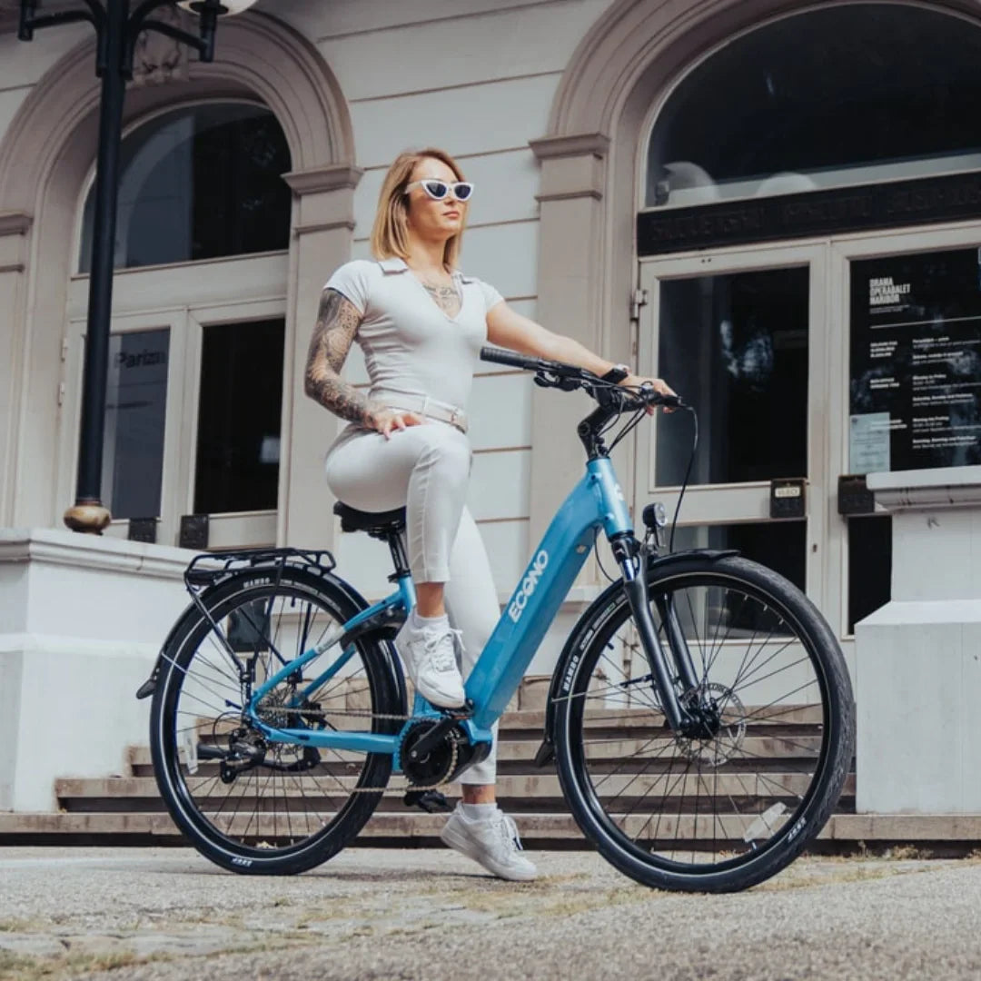 The product pictured is an electric bike, brand Econo, model EEC4LK. The image shows a woman sitting on a blue electric bike, dressed in a light outfit and sunglasses. The bike has wide tires and is positioned in front of an elegant building, giving the impression of an urban setting.