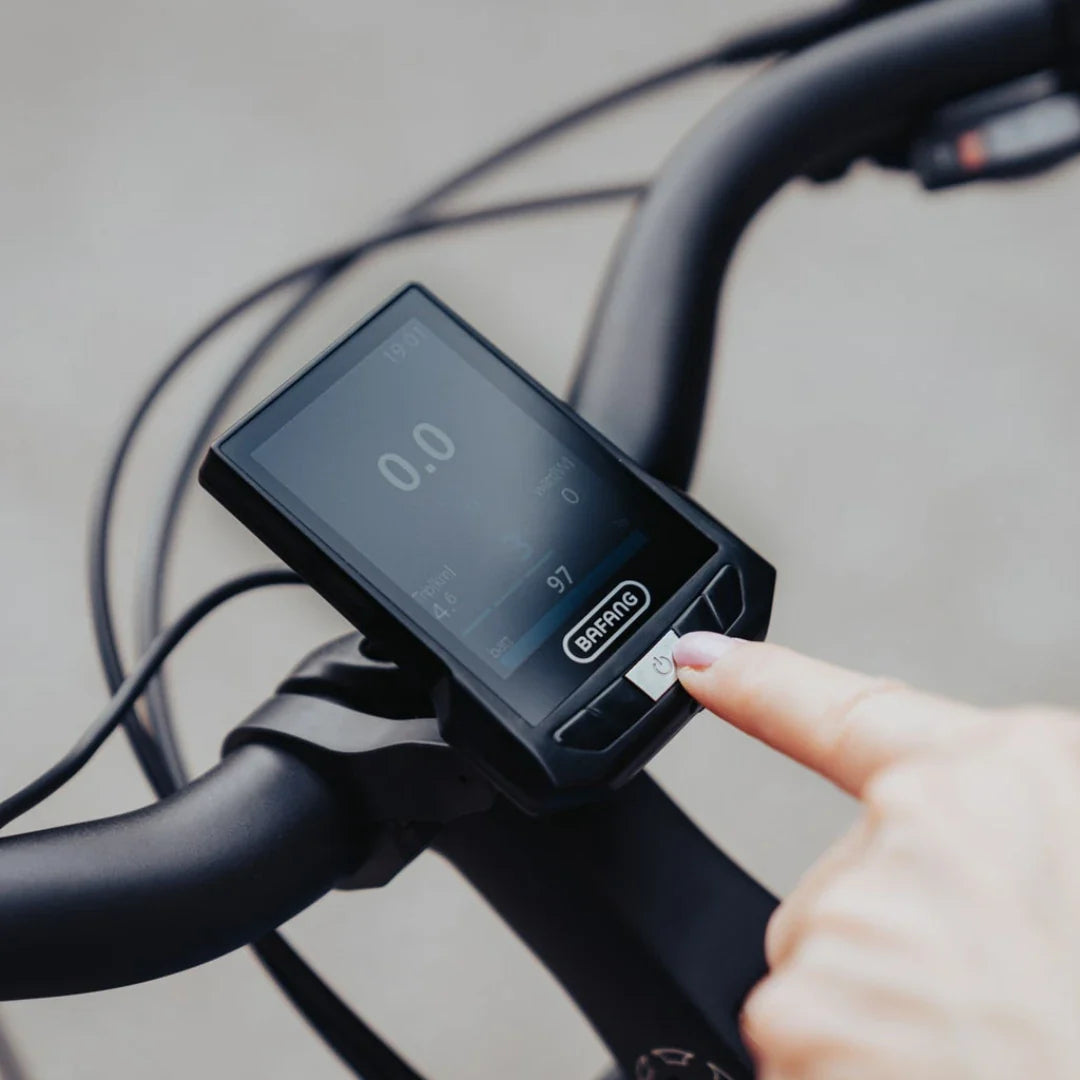 The product in the image is an Electric bike, model Econo Mare City. The image shows a display that indicates various data, such as speed and battery status. The user is pressing a button on the display, which features the Bafang brand, suggesting the use of an electric drive system from this brand. The bike has ergonomically designed handlebars and a modern design.
