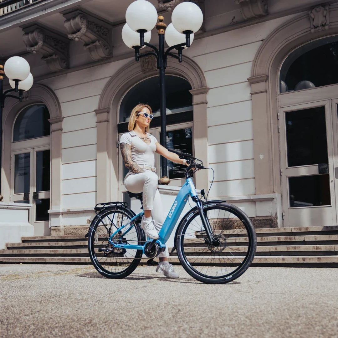The image shows an Electric Bike Econo Mare City. The bike is light blue and has a sporty design. A woman in a light dress is sitting on the bike in front of an elegant building with stairs and lamps. Large windows are visible in the background, adding to the urban ambiance of the photo.