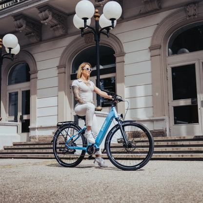 The image shows an Electric Bike Econo Mare City. The bike is light blue and has a sporty design. A woman in a light dress is sitting on the bike in front of an elegant building with stairs and lamps. Large windows are visible in the background, adding to the urban ambiance of the photo.