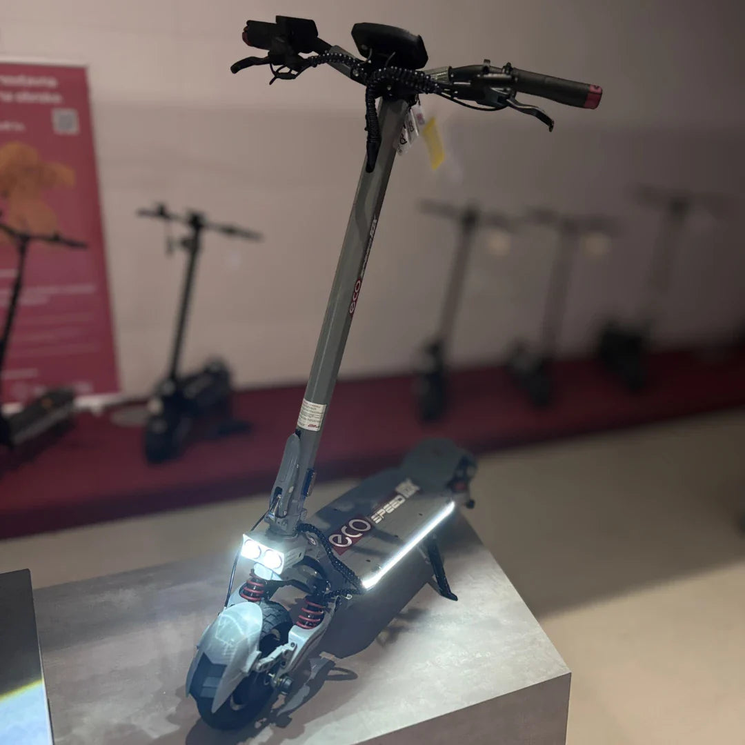 Izdelek na sliki je električni skiro, model Eco Speed 10X V2. Na sliki je prikazan skiro v modernem dizajnu, z vidnimi svetilkami spredaj in robustno konstrukcijo. Na ročaju so nameščene ročice za upravljanje, medtem ko je v ozadju mogoče videti še več skirojev, kar nakazuje na razstavo ali trgovino.