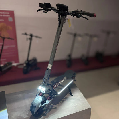 Izdelek na sliki je električni skiro, model Eco Speed 10X V2. Na sliki je prikazan skiro v modernem dizajnu, z vidnimi svetilkami spredaj in robustno konstrukcijo. Na ročaju so nameščene ročice za upravljanje, medtem ko je v ozadju mogoče videti še več skirojev, kar nakazuje na razstavo ali trgovino.