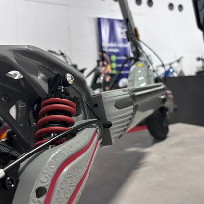 Na sliki je električni skiro, model Eco Speed 10X V2. Vidna je podrobnost vzmetenja s rdečimi vzmetmi ter del okvirja skiroja. Ozadje slike prikazuje druge električne naprave, kar nakazuje, da je skiro del razstave ali trgovine.