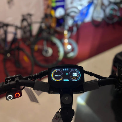 Na sliki je električni skiro, model Eco Speed 10X V2. Vidna je krmilo skiroja s digitalnim zaslonom, ki prikazuje hitrost in način vožnje. V ozadju so vidni tudi drugi kolesi, kar nakazuje, da je slika posneta v trgovini ali na razstavi koles.