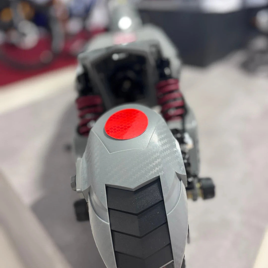 Izdelek na sliki je električni skiro, model Eco Speed 10X V2. Na sliki je vidna zadnja stran skiroja, kjer so prikazani amortizerji v rdeči barvi ter del zadnjega kolesa s profilom pnevmatike. Vidna je tudi odsevna oznaka v rdeči barvi, kar povečuje varnost pri vožnji. Skupna zasnova skiroja je moderna in funkcionalna.