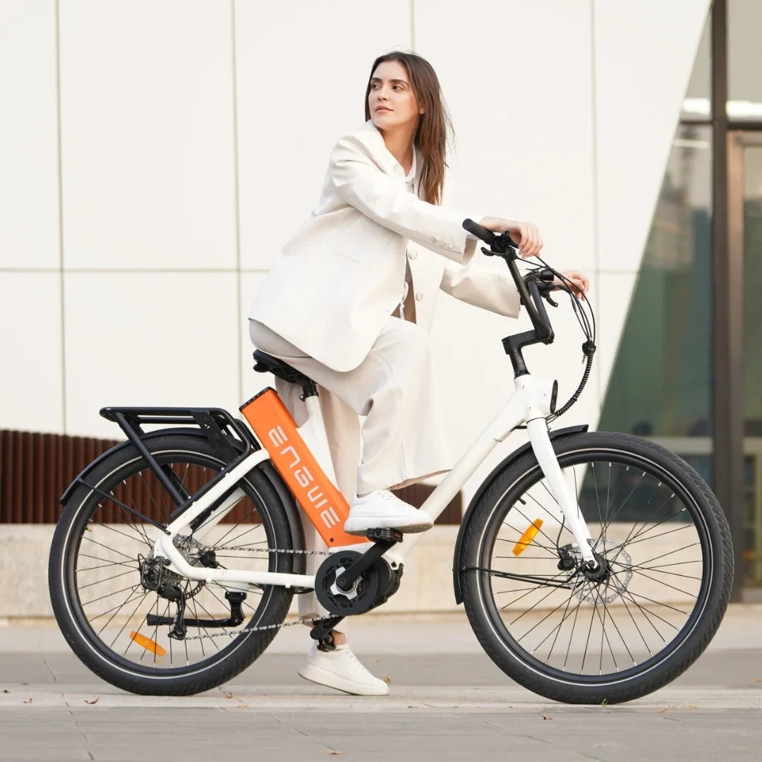 The product is an Electric bike, Engwe P275 ST model. The image shows a woman sitting on the bike, dressed in light-colored clothing. The bike is white and features a distinctive orange battery on the frame and a luggage rack. Modern architecture in the background gives the image a contemporary look.