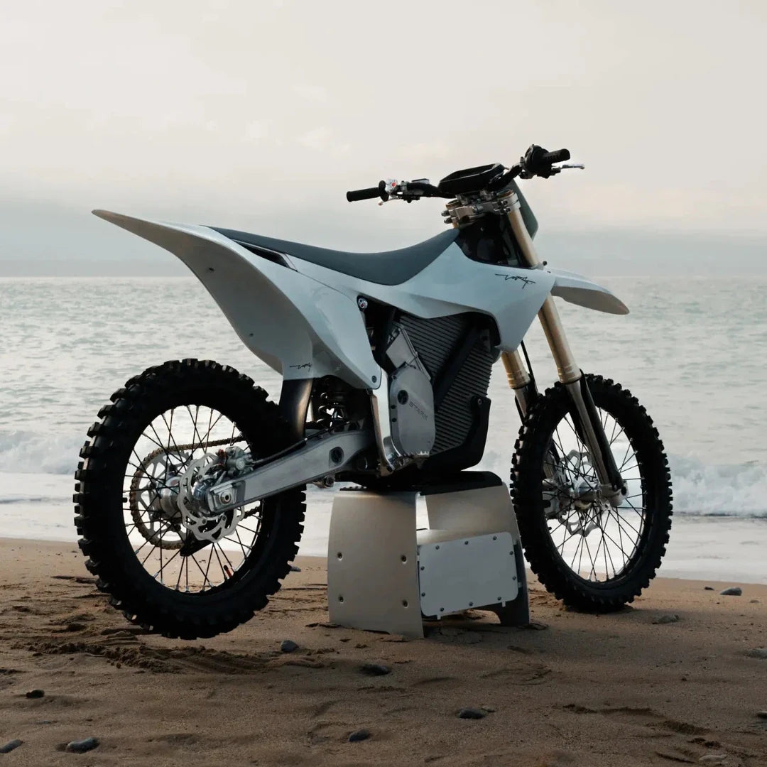 Electric Motocross Motorcycle Stark Varg parked on a sandy beach with ocean waves in the background.