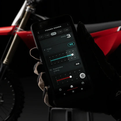 A gloved hand holds a smartphone displaying settings for the Stark Varg electric motocross motorcycle.
