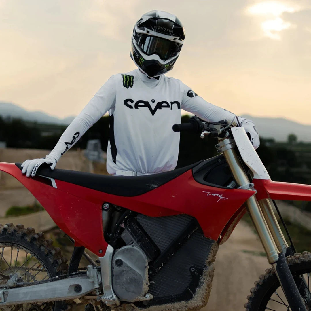 A rider stands next to a Stark Varg electric motocross bike with a sunset in the background.