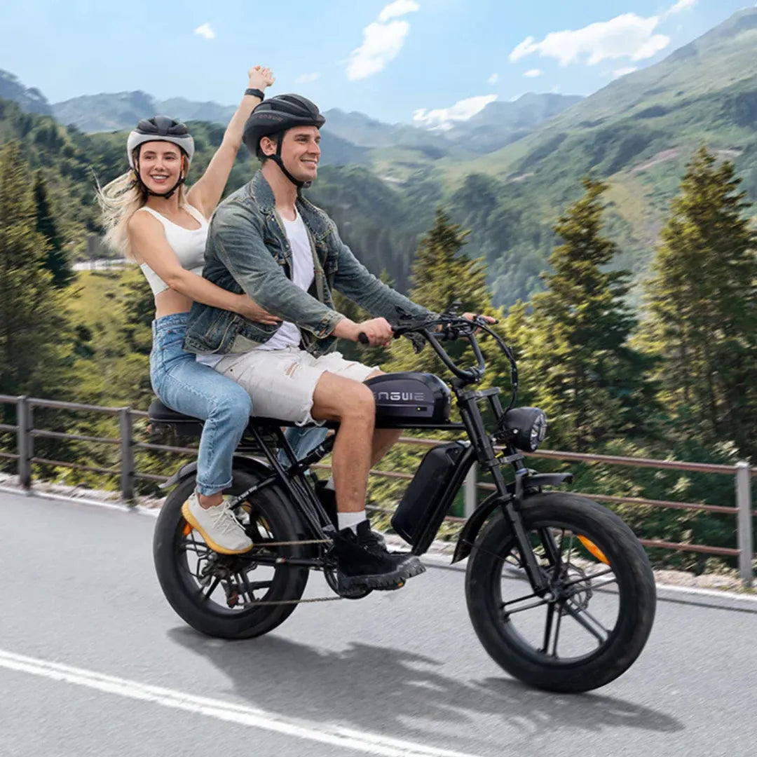 The product in the image is an electric bike from the brand Engwe, model M1. Two people are visible in the picture, riding the bike on a scenic road next to a wooded area. The cycling enthusiasts are dressed in sportswear and wearing helmets. The bike has wider tires and a modern design, suggesting a comfortable ride.