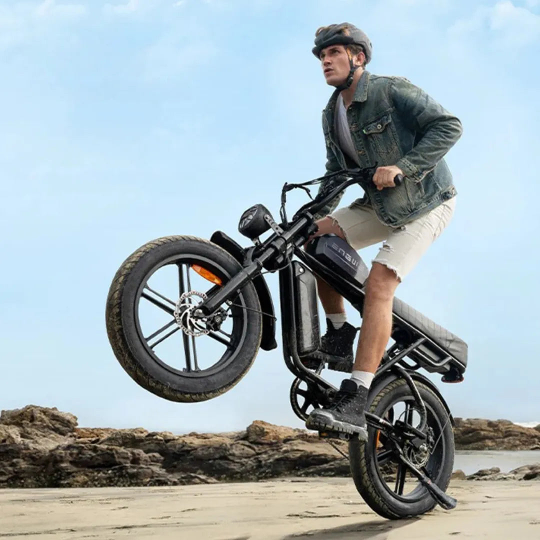 The image shows an Electric bike from the Engwe brand, model M1. It depicts a man performing a trick by lifting the front wheel off the ground while riding on a beach. The bike has a robust design, wide wheels, and is equipped with an electric motor and battery, allowing for comfortable riding on various terrains. The man is wearing a helmet, a denim jacket, and shorts.