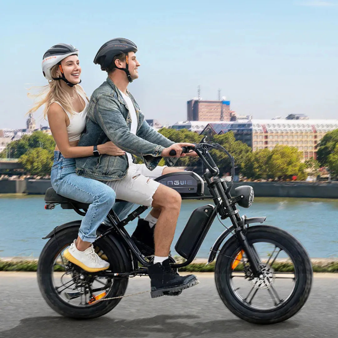 The product in the image is an Electric bike, model Engwe M1. Two riders, a man and a woman wearing helmets, are visible on the bike. The bike has wide tires and is designed for comfortable riding. In the background, there's a scenic view of a river and city architecture, creating a pleasant atmosphere.