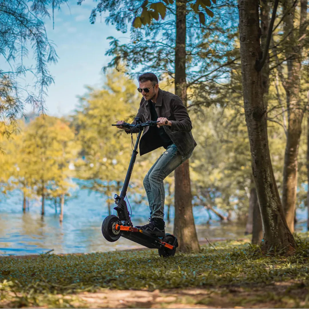 The image shows an Electric Scooter, model Joyor S10 S-Z. A man is seen riding the scooter in a natural environment, by the water and among trees. He is wearing sunglasses and appears to be enjoying the ride. The scooter is black with orange accents, giving it a sporty look.