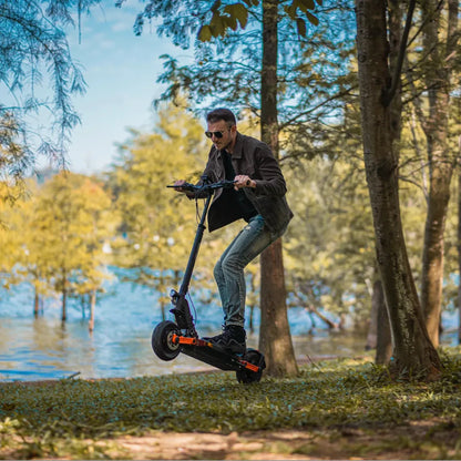 The image shows an Electric Scooter, model Joyor S10 S-Z. A man is seen riding the scooter in a natural environment, by the water and among trees. He is wearing sunglasses and appears to be enjoying the ride. The scooter is black with orange accents, giving it a sporty look.