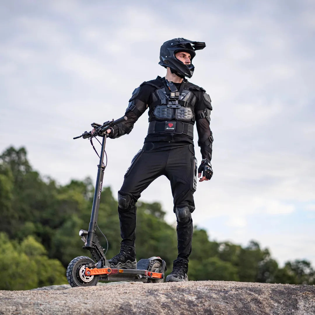 The image shows an Electric Scooter Joyor S10-S Z. We see a rider, dressed in protective gear, standing on the scooter on a rocky surface. The surroundings are natural, with greenery in the background, suggesting that the scooter is suitable for off-road riding. The rider is wearing a helmet and protective clothing, which emphasizes safety during the ride.