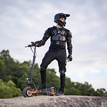 The image shows an Electric Scooter Joyor S10-S Z. We see a rider, dressed in protective gear, standing on the scooter on a rocky surface. The surroundings are natural, with greenery in the background, suggesting that the scooter is suitable for off-road riding. The rider is wearing a helmet and protective clothing, which emphasizes safety during the ride.