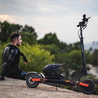 The image shows an Electric Scooter, model Joyor S10 S-Z. The product is equipped with robust wheels and a sporty design, indicating its performance capabilities. A man wearing protective gear is seated next to the scooter, suggesting its intended use for thrill rides or sports activities. Green tree canopies and a part of the city landscape are visible in the background.
