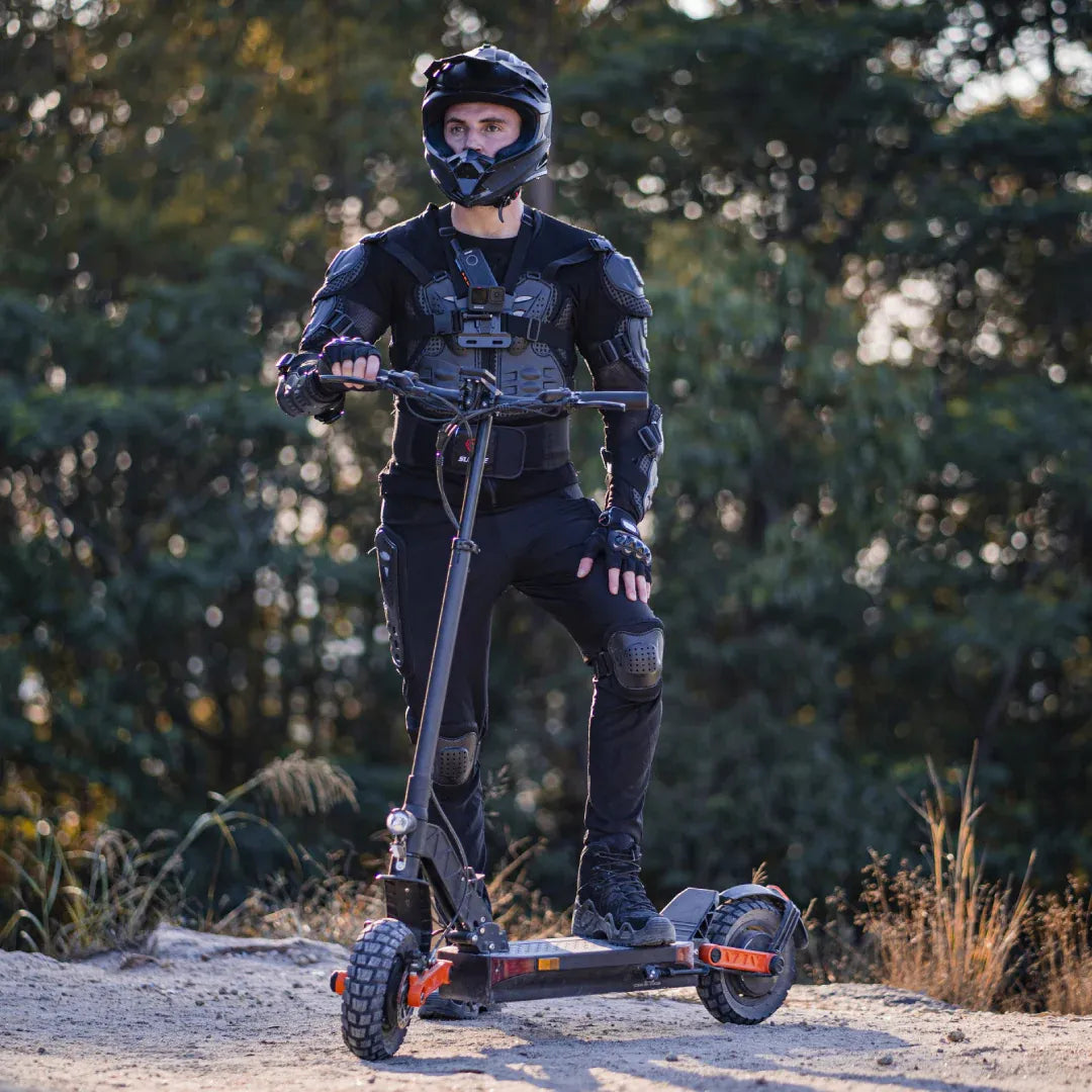 The product in the image is an Electric Scooter, model Joyor S10 S-Z. The image shows a man, dressed in black protective gear and a helmet, standing on the scooter outdoors. The surroundings are natural, with trees in the background. The scooter's design is robust, suggesting its suitability for various terrains.