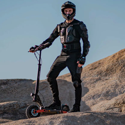 The image shows an Electric Scooter, model Joyor S10 S-Z. The person standing on the scooter is wearing protective gear, including a helmet and protective clothing, indicating preparation for riding in more demanding terrains. The carefully chosen rocky terrain further highlights the scooter's capabilities.
