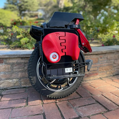 The product in the image is an electric unicycle, model KingSong S22 Pro. The image shows a folded unicycle in black and red. Large wheels with a robust tread are visible, as well as the section where the controls and battery are located. The unicycle is placed on a stony surface, with plants visible in the background, giving a sense of an outdoor environment.