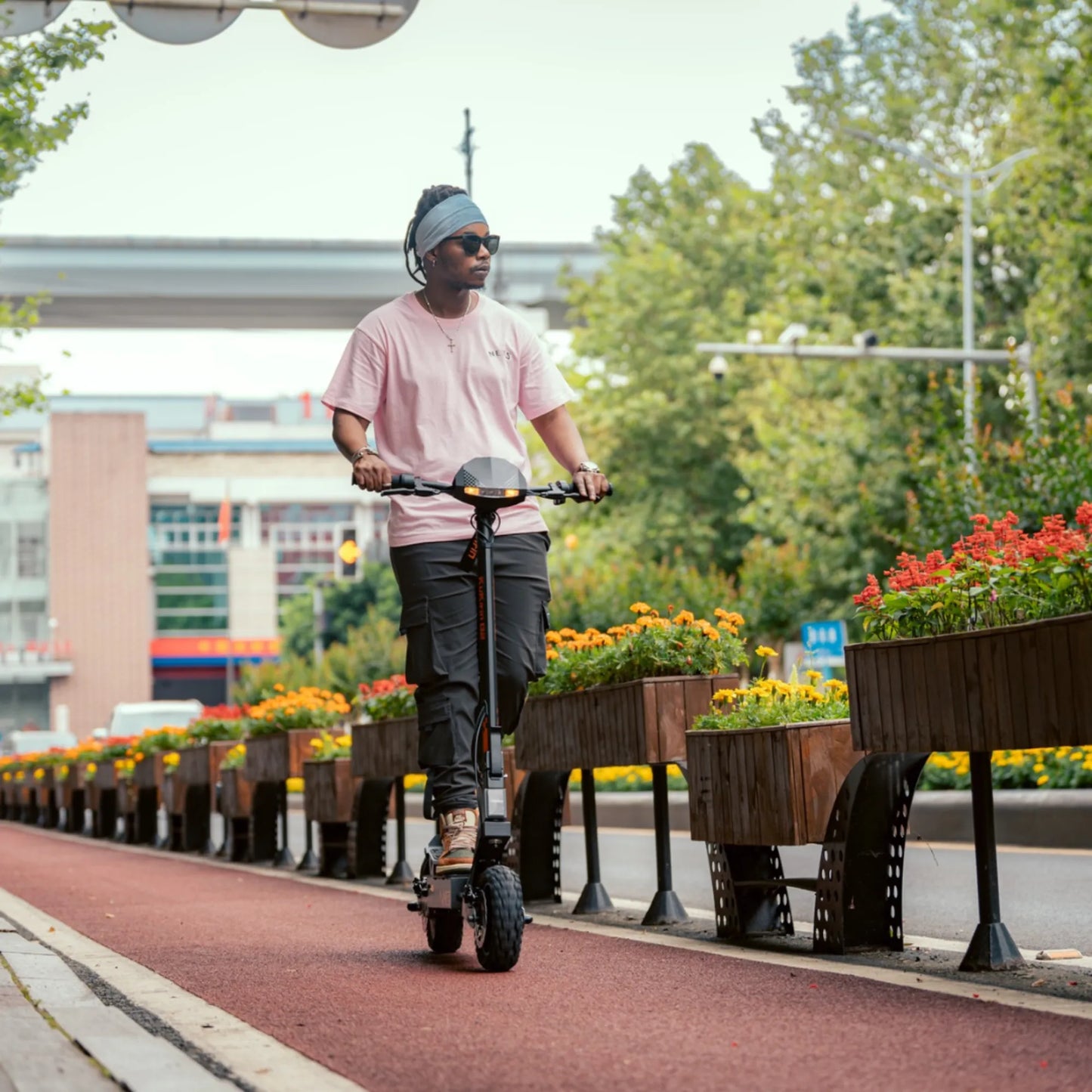 The product in the image is an Electric Scooter KuKirin G2. The image shows a person riding the scooter on a paved path next to flower beds. The person is wearing a pink T-shirt and sunglasses, and the surroundings are green and pleasant, creating a relaxed atmosphere.