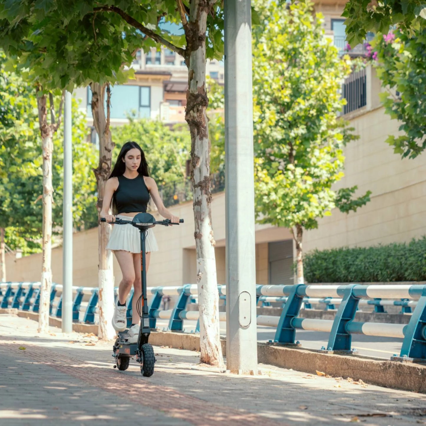 The image shows an Electric Scooter KuKirin G2. A person on the scooter is riding along a paved path next to trees, indicating a pleasant environment for riding. The product features robust tires, allowing for a comfortable ride on various surfaces.
