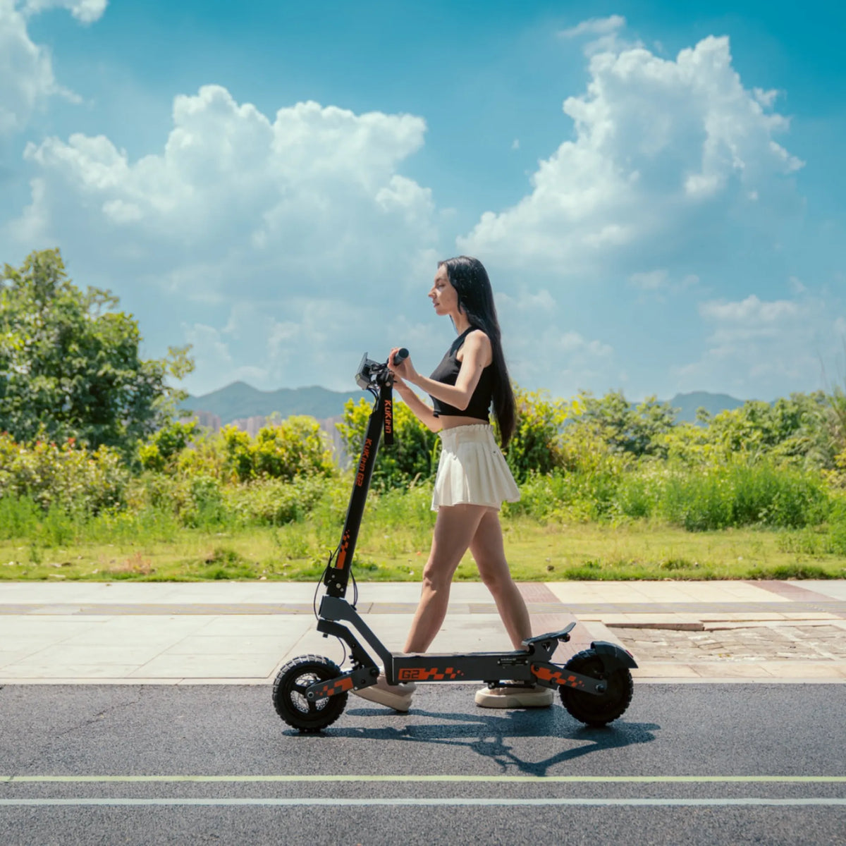 The image shows an Electric Scooter KuKirin G2. We see a young woman riding the scooter on a wide path alongside a green environment. In the background, there are trees and hills, and the sky is clear with a few clouds. Overall, the picture displays a modern and elegant electric scooter in use.
