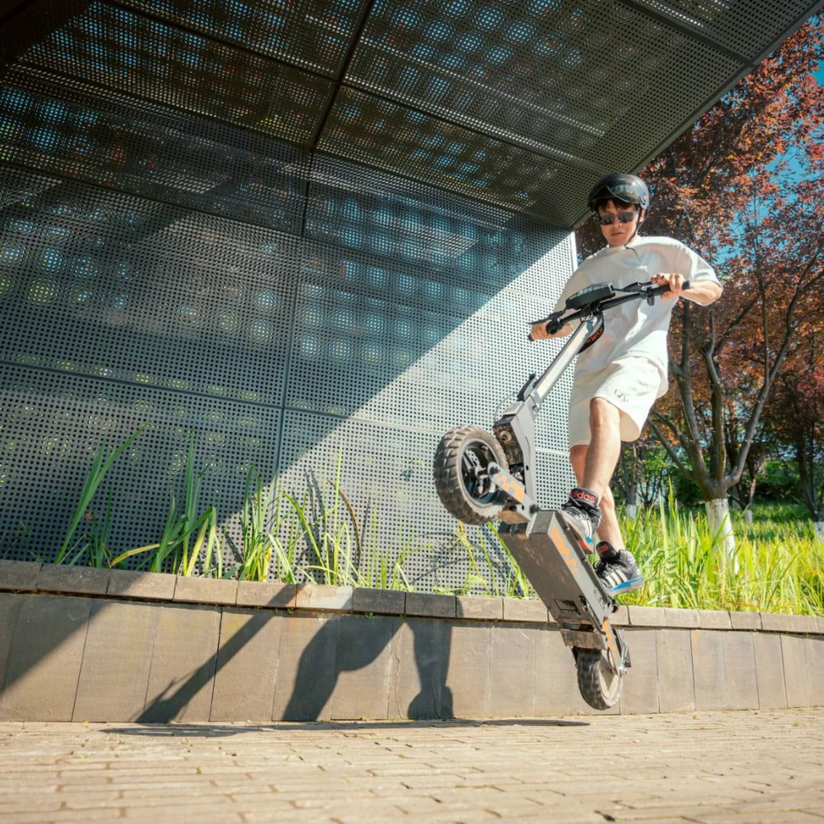 The image shows an Electric Scooter KuKirin G2. The user is performing an acrobatics, lifting the front wheel off the ground while standing on the pavement next to greenery. It is equipped with robust tires, indicating its suitability for various terrains. In the background, a modern architectural element adds dynamism to the overall picture.