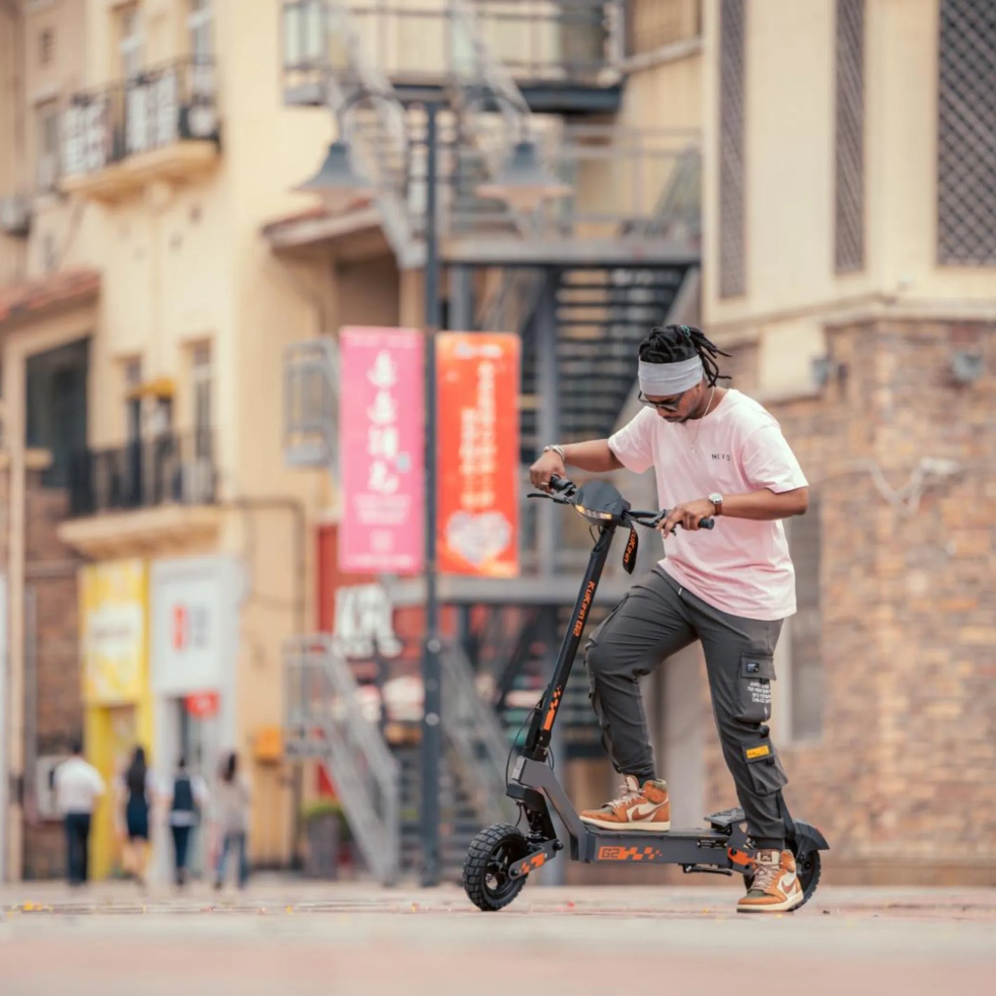 The product in the image is an Electric Scooter, model KuKirin G2. The image shows a person riding the scooter in an urban environment. It is equipped with robust wheels and has a modern design, suggesting a comfortable ride. Buildings and city infrastructure are visible in the background, creating a dynamic urban atmosphere.