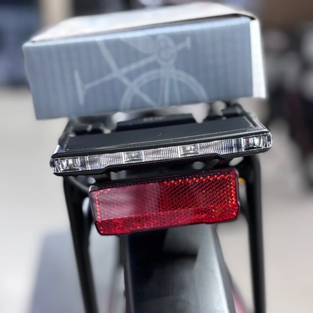 The product in the image is a Malaguti electric bike, model Bolonina. The rear light and reflector, mounted on the bike's frame, are visible in the picture. A box, likely containing accessories or instructions, is also in the background. Lights and a reflector are important for safe riding in the dark.