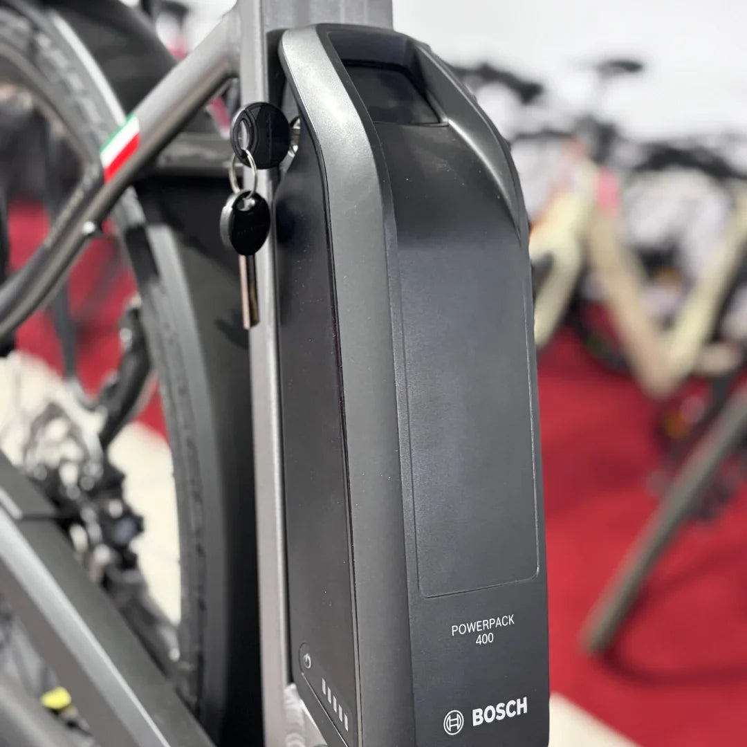 The product is an Electric bike of the brand Malaguti, model Bolonina. The image shows a Bosch Powerpack 400 battery mounted on the bike's frame. The battery has a key and a charge indicator, suggesting its functionality and ease of use. Other bikes are also visible in the background, creating the impression of an exhibition.