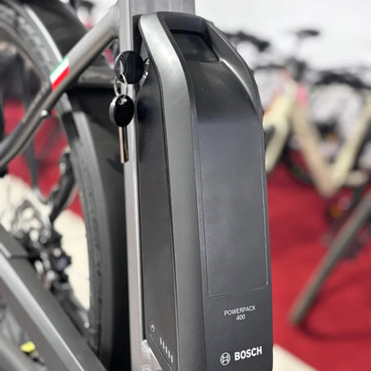 The product is an Electric bike of the brand Malaguti, model Bolonina. The image shows a Bosch Powerpack 400 battery mounted on the bike's frame. The battery has a key and a charge indicator, suggesting its functionality and ease of use. Other bikes are also visible in the background, creating the impression of an exhibition.