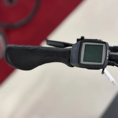 The image shows an electric bike from the Malaguti brand, model Bolonina. Visible are the handlebars with black grips and a display showing information about the bike's operation. The display has buttons for adjustment, indicating functionalities such as speed selection or riding mode. The background of the image is blurred, but the space appears tidy and suitable for showcasing the bike.