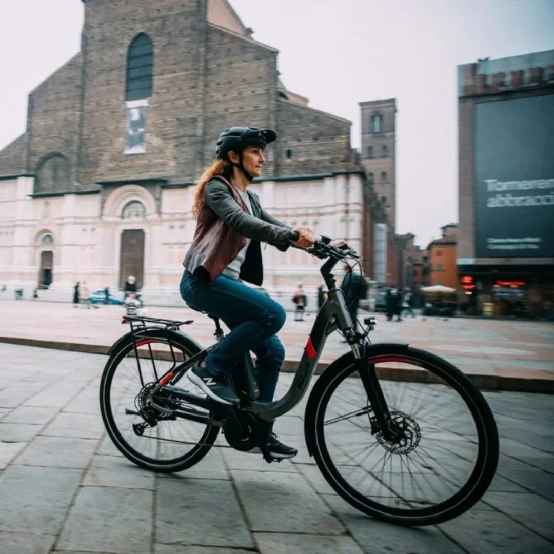 The image shows an Electric bike by Malaguti, model Bolonina. It depicts a woman riding the bike through a city square surrounded by architectural landmarks. The bike features a modern design and is equipped with an electric motor and a comfortable seat, ensuring a pleasant ride. She is dressed in comfortable clothing and a helmet, indicating an awareness of safety while riding.