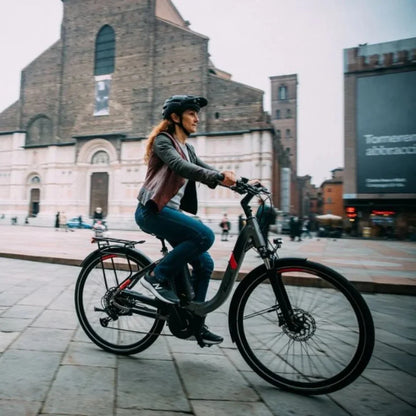 The image shows an Electric bike by Malaguti, model Bolonina. It depicts a woman riding the bike through a city square surrounded by architectural landmarks. The bike features a modern design and is equipped with an electric motor and a comfortable seat, ensuring a pleasant ride. She is dressed in comfortable clothing and a helmet, indicating an awareness of safety while riding.