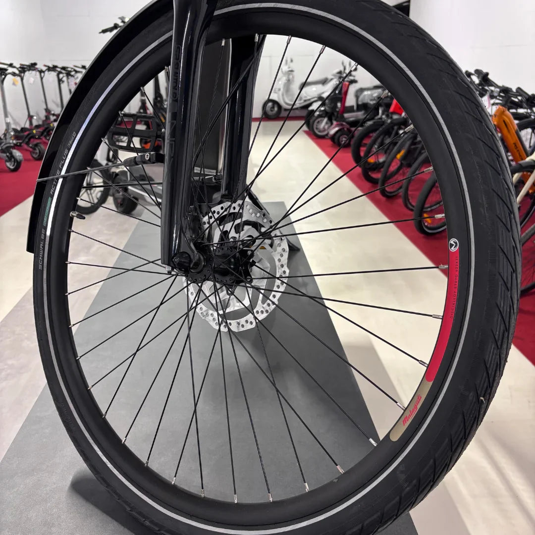 The product in the image is an electric bike from the brand Malaguti, model Bolonina. The image shows the front wheel, which includes disc brakes and a tire with a distinctive tread. In the background, other electric scooters and bikes are visible, suggesting it is a point of sale or an exhibition.