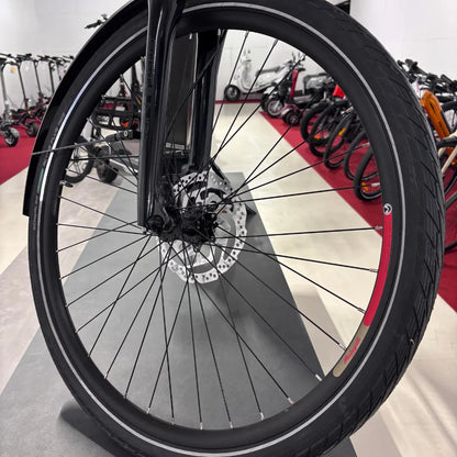 The product in the image is an electric bike from the brand Malaguti, model Bolonina. The image shows the front wheel, which includes disc brakes and a tire with a distinctive tread. In the background, other electric scooters and bikes are visible, suggesting it is a point of sale or an exhibition.