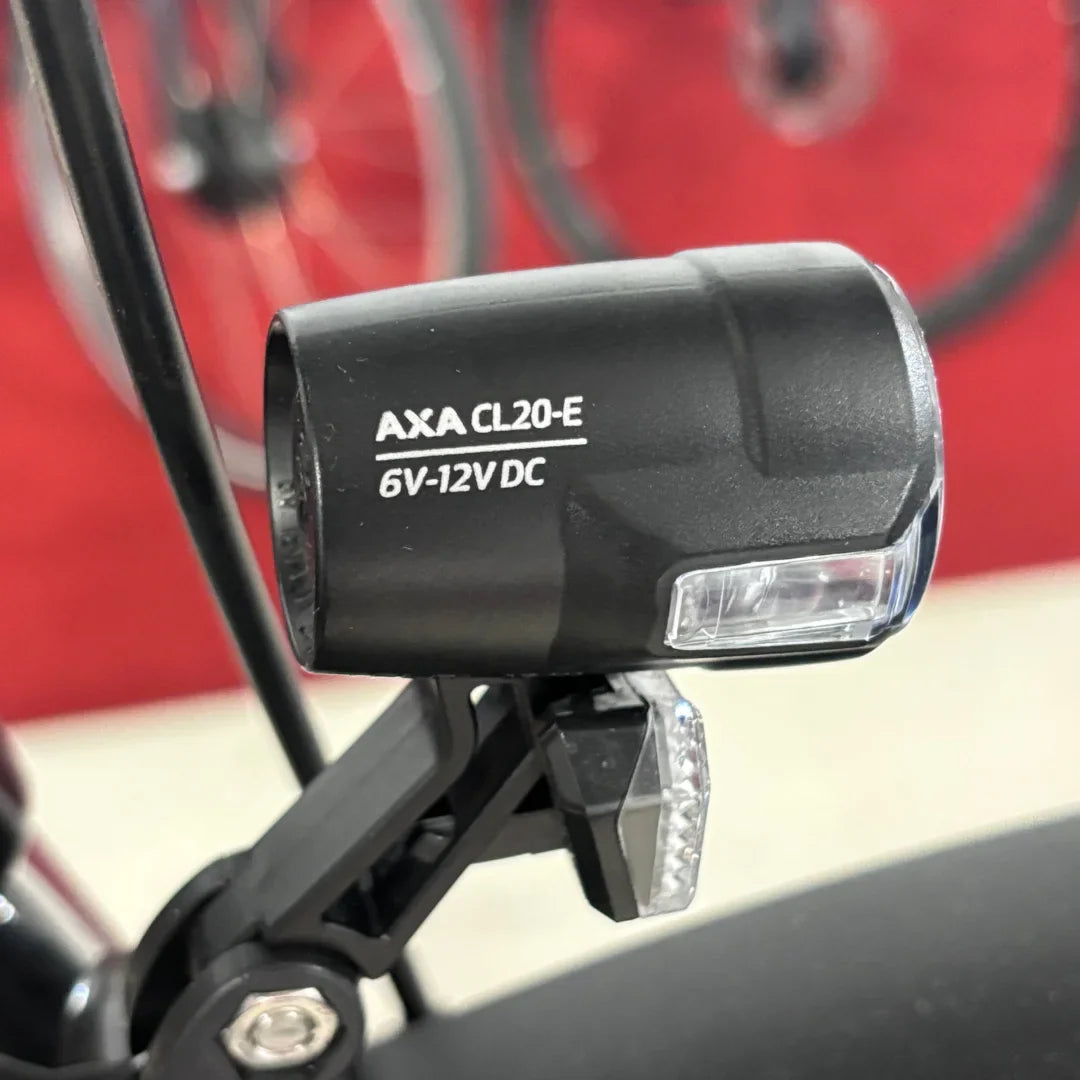 The image displays a light for an electric bike, brand AXA, model CL20-E, operating at 6V-12V DC. The light is mounted on the bike frame and is designed to improve visibility while riding. The bike's wheels are partially visible in the background, indicating it is an electric bike.