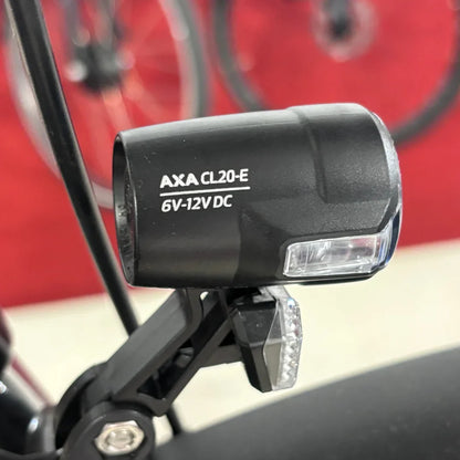 The image displays a light for an electric bike, brand AXA, model CL20-E, operating at 6V-12V DC. The light is mounted on the bike frame and is designed to improve visibility while riding. The bike's wheels are partially visible in the background, indicating it is an electric bike.