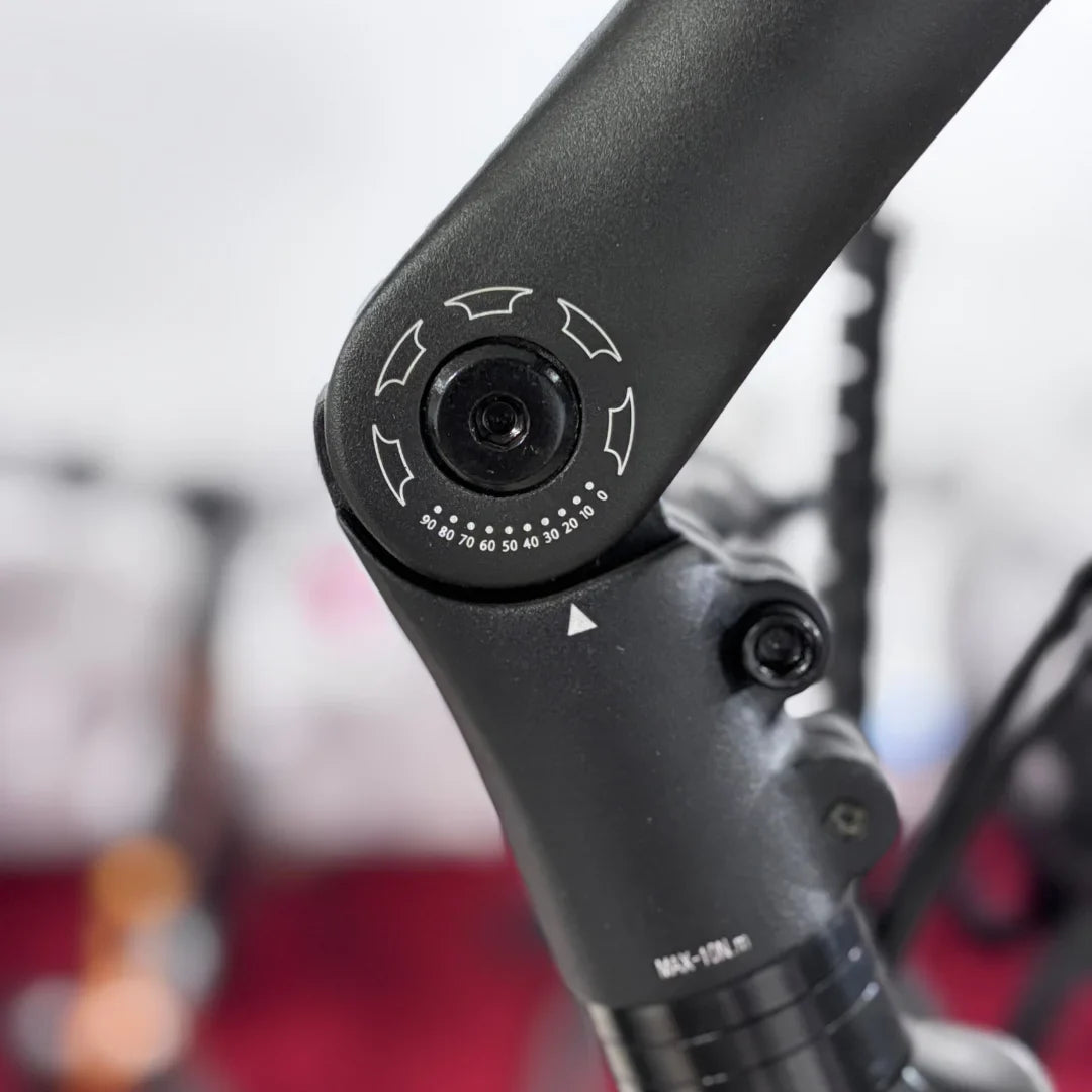 The image shows a Malaguti electric bike, model Bolonina. A detail on the frame is visible, where an adjustment button is located, allowing for the customization of various bike functions. The button has marked values and directions, indicating adjustable settings. The background of the image is blurred, emphasizing the bike.