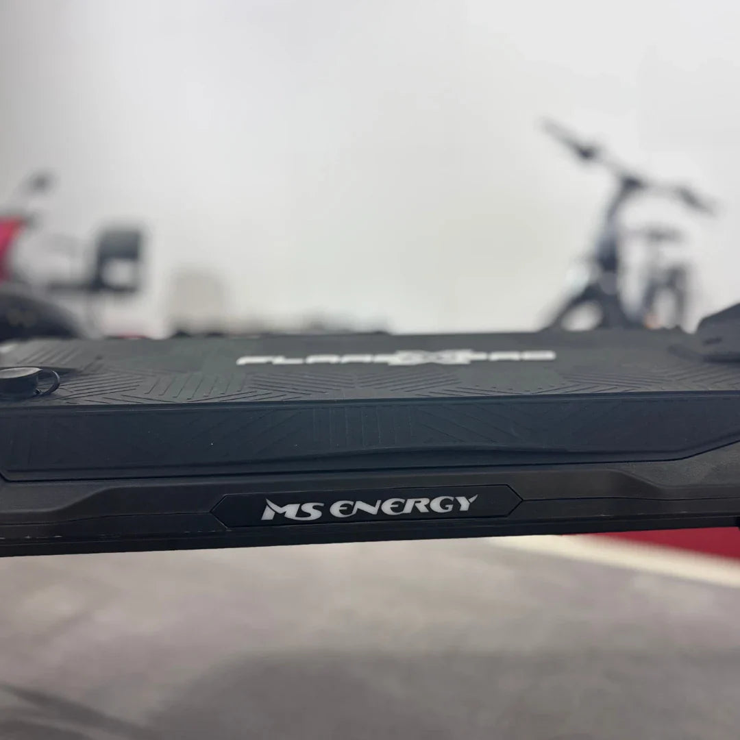Na sliki je električni skiro, model MS Energy Flare X Pro. Vidna je platforma skiroja z izrazitim vzorcem, ki zagotavlja dober oprijem, ter logotip znamke MS Energy. V ozadju so delno vidni drugi kolesarski izdelki, kar nakazuje, da je slika posneta v okolju, kjer se prodajajo električni prevozi.
