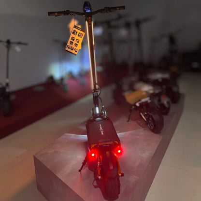 Izdelek na sliki je električni skiro, model MS Energy Flare X Pro. Na sliki je vidna zadnja stran skiroja, ki ima močne rdeče luči, kar nakazuje na njegovo funkcionalnost pri vožnji v temi. Na krmilu je pritrjen informativni listič, ki verjetno vsebuje specifikacije ali navodila za uporabo. V ozadju so vidni tudi drugi električni skiroji, kar nakazuje na razstavo ali prodajno mesto.