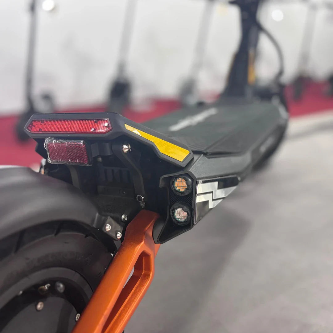 Izdelek je električni skiro, model MS Energy Flare X Pro. Na sliki je vidna zadnja stran skiroja, ki vključuje svetlobne elemente, kot so rdeča in rumena luč ter reflektor. Ohišje skiroja je črno z oranžnimi detajli, kar mu daje dinamičen videz. V ozadju so vidni tudi drugi skiroji, kar nakazuje na okolje, kjer so predstavljeni.